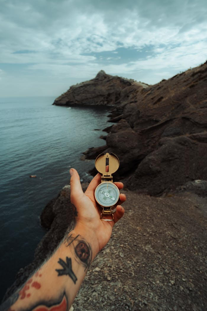 A tattooed hand holding a compass by the sea against a cliff background, perfect for adventure themes.