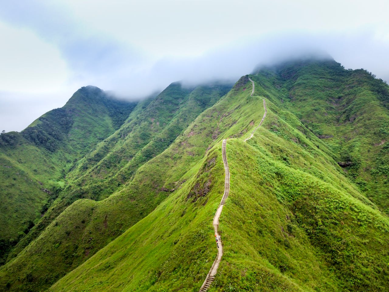 A dramatic drone view of a lush green mountainous landscape with a winding footpath under clouds.